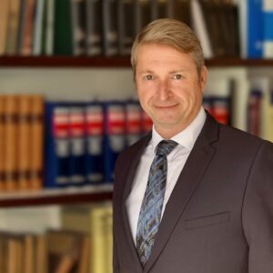 Ein lächelnder Mann im Anzug vor einem Bücherregal mit bunten Büchern